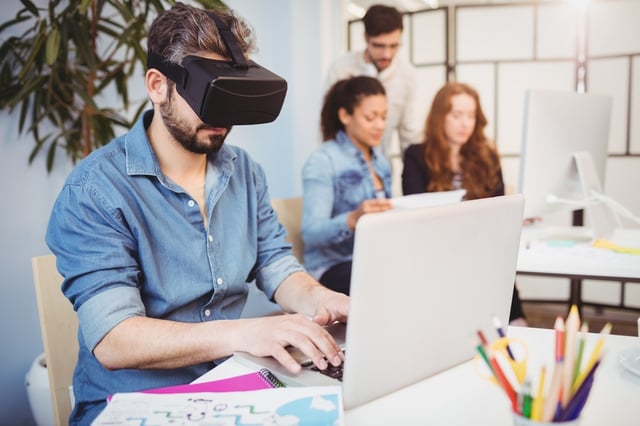 Businessman wearing virtual reality headset while using laptop against colleagues in creative office.jpeg